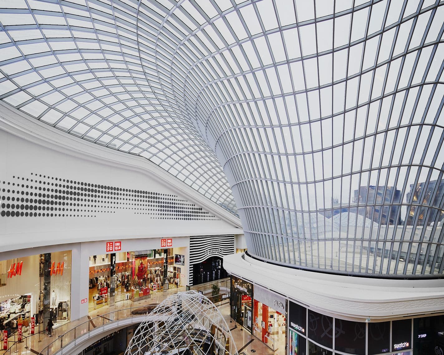Interior view of a shopping centre featuring a sweeping glass roof and modern architectural design. Shops such as H&M and Uniqlo line the upper level, with pathways for shoppers below.