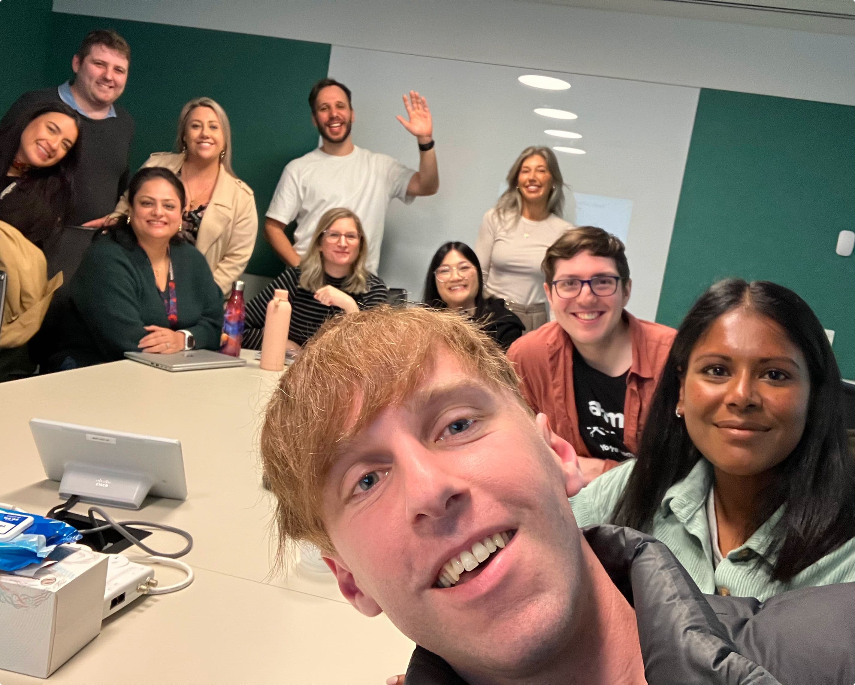 A group of twelve people gathered around a meeting table in a modern office, smiling and posing for a selfie. The group appears friendly and engaged, with one person in the foreground taking the photo and others waving or smiling in the background.