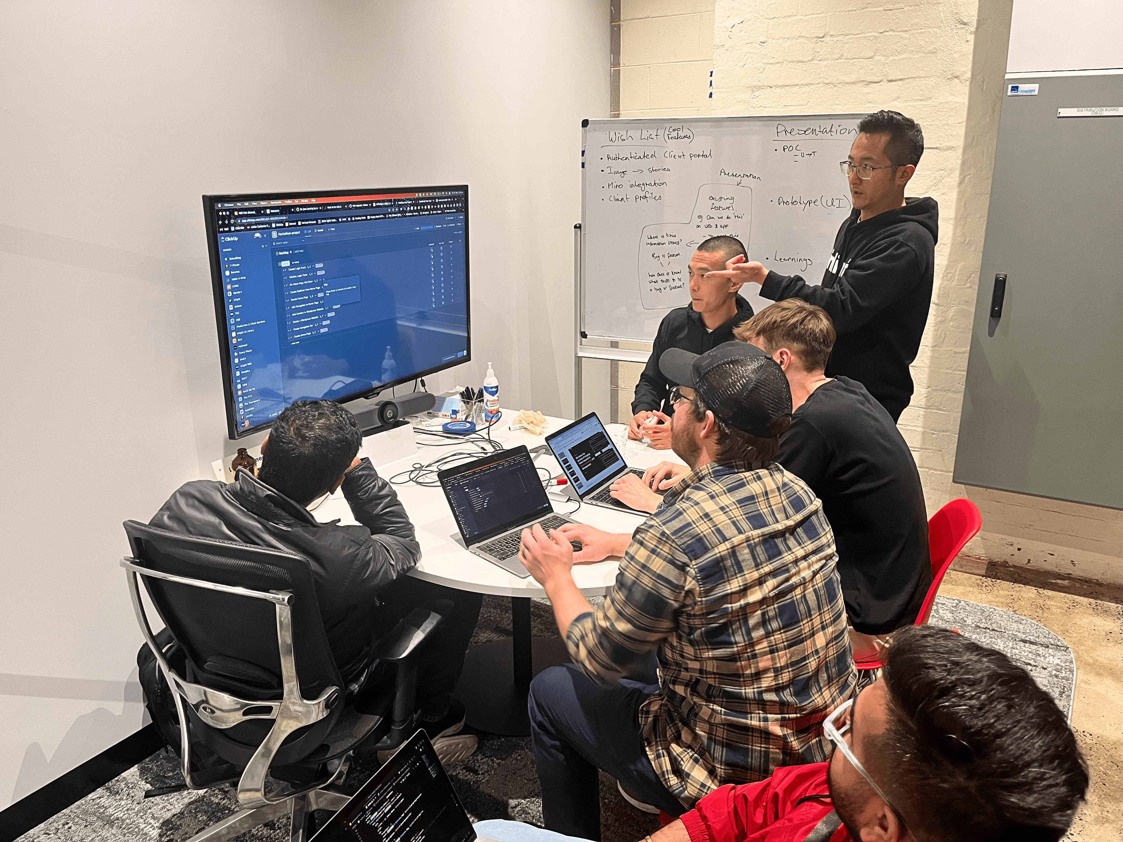 A small team sits around a table in a collaborative workspace, discussing code displayed on a large screen. One person stands near a whiteboard covered with notes, guiding the conversation while others work on laptops.