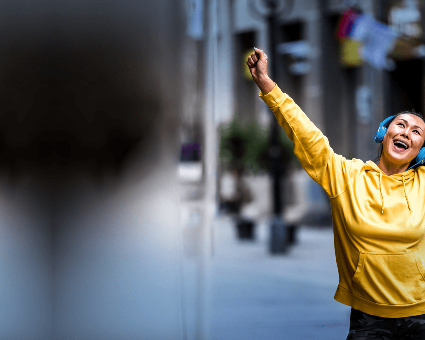 A full-width image of a woman in a yellow hoodie raising her fist triumphantly while listening to music with bright blue headphones, walking outdoors in an urban setting with blurred buildings in the background.