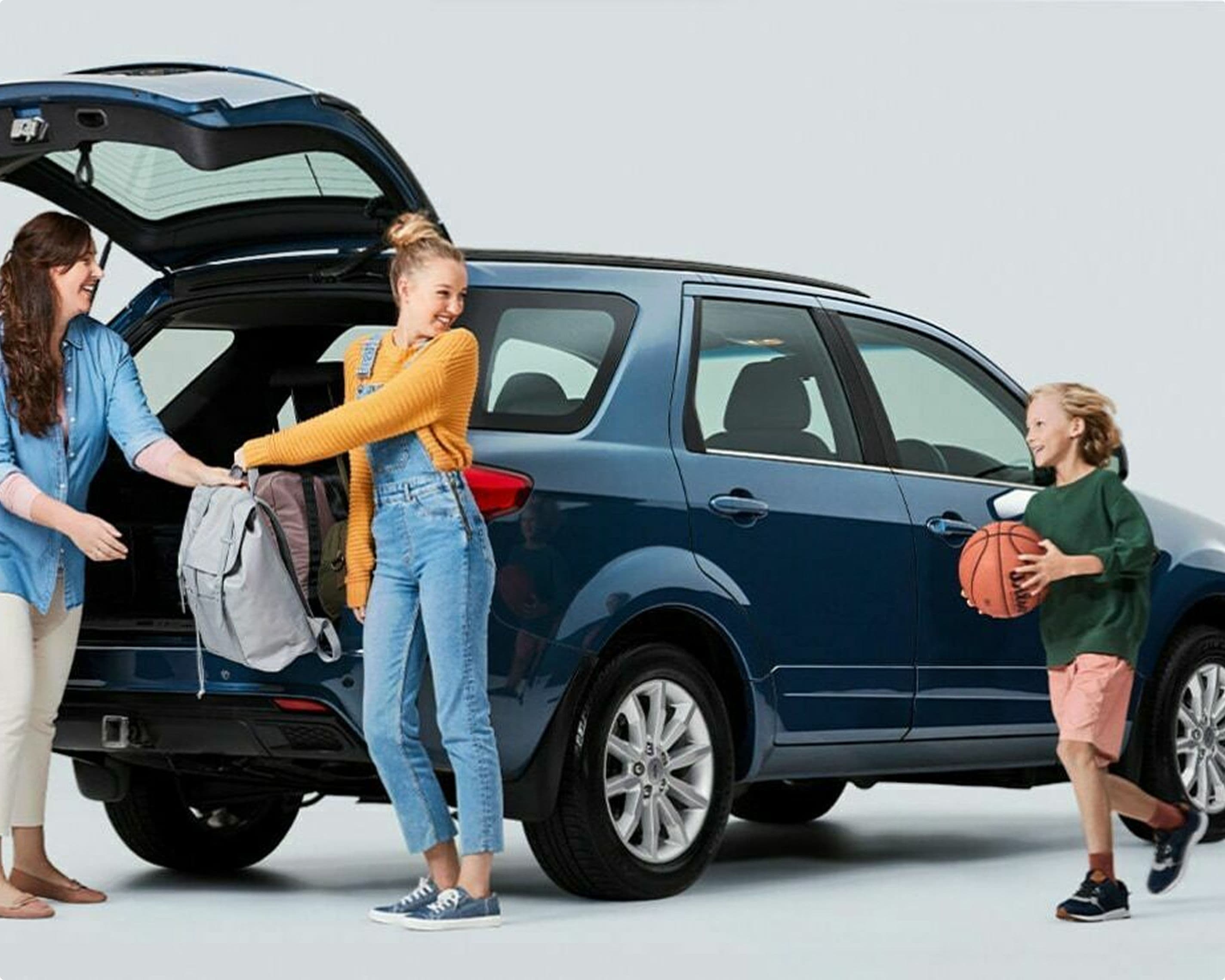 A family is unloading items from the back of a blue SUV. An adult hands a backpack to a teenager, while a child holding a basketball stands by the car. The scene suggests a family outing or after-school activity.