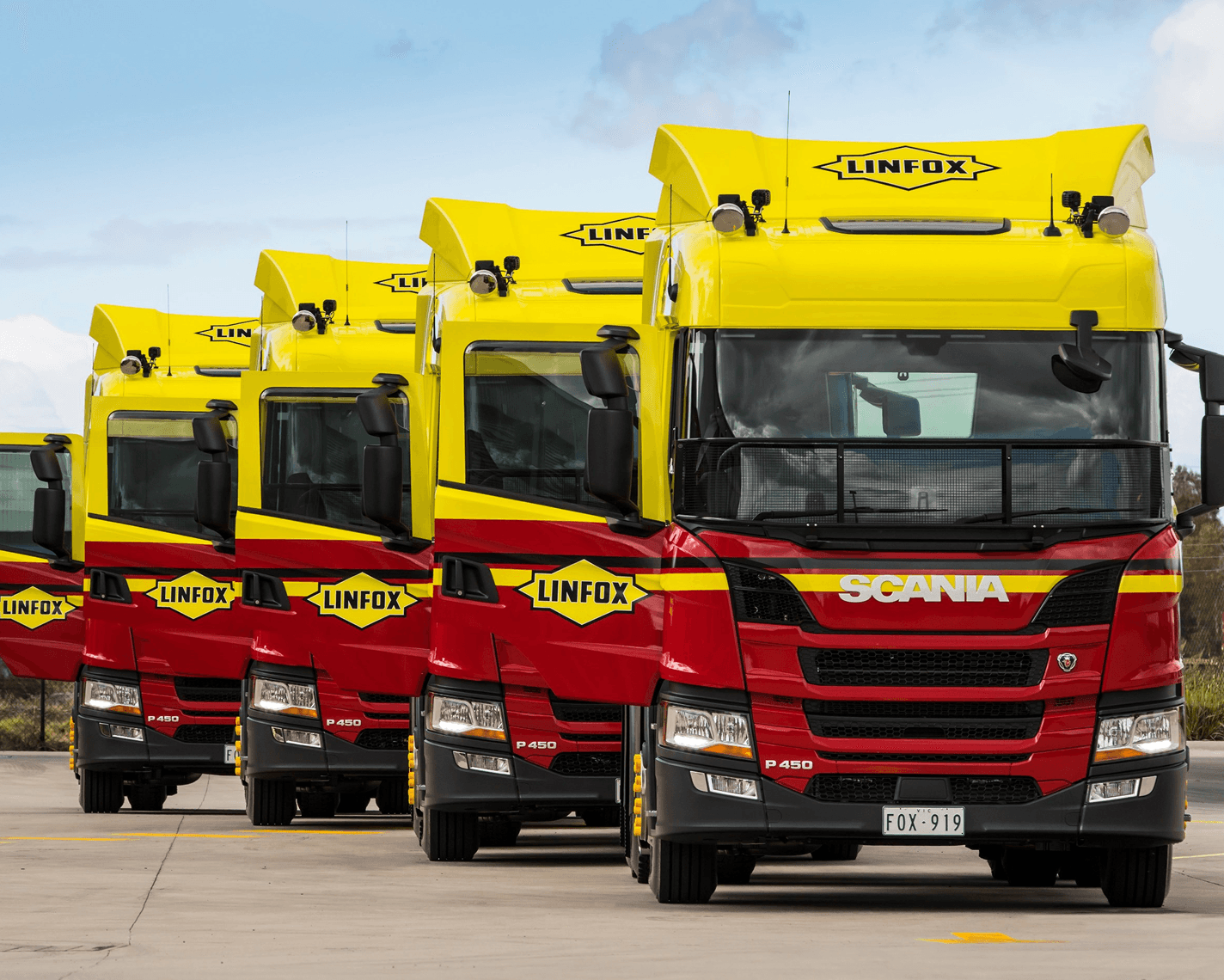 A row of large, modern Linfox trucks is parked outdoors. The trucks are painted in the signature Linfox colours of yellow and red, with bold Linfox logos visible above the windscreen and on the side. The trucks are Scania brand, with the model “P450” displayed. The sky is partly cloudy, and the trucks are lined up on a concrete surface.
