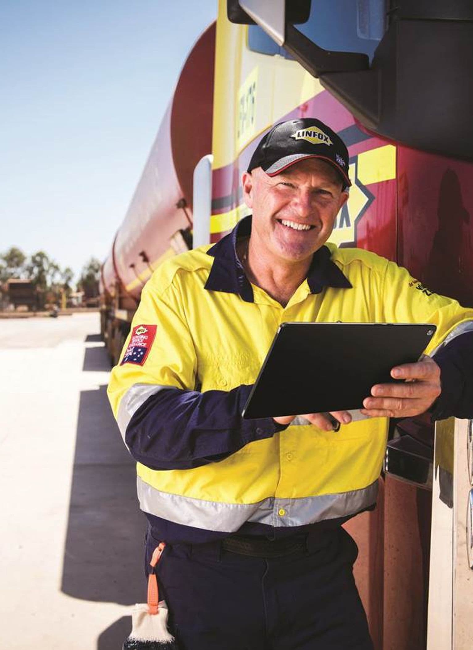 A Linfox worker is wearing a high-visibility yellow and navy uniform, using a tablet while leaning against a Linfox-branded truck. The worker’s cap and clothing feature the Linfox logo, and the setting appears to be an outdoor logistics or depot area, with more trucks and trees in the background.